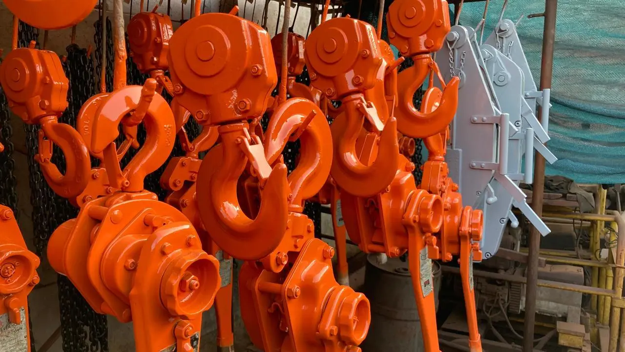 Tecles mecánicos de cadena recién pintados, en color naranja brillante, colgados en fila junto a otros en color gris, listos para su uso o almacenamiento.